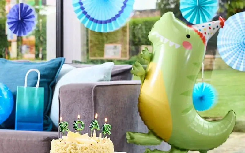 Dinosaur-themed birthday party display with a birthday cake, dinosaur balloon, blue decorations, gift bags, and a lit number 4 decoration.